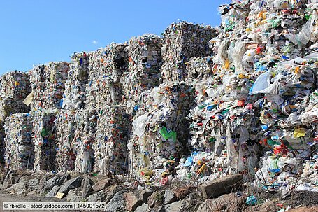 Photo of baled waste plastics with mixed colours stacked outdoors in a yard seen against the sky. 