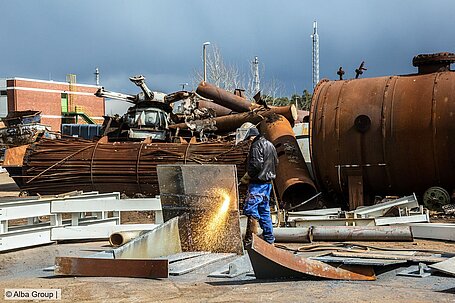 Photo of a scrapyard with a worker cutting a metal sheet using a torch cutting tool.