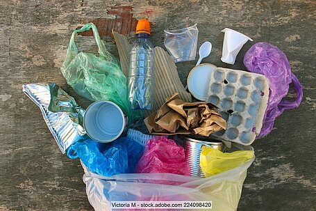 Various household packaging items and plastic bags on a flat surface, including an aluminium tray, several tins, a yoghurt pot, an egg carton, crumpled paper, a plastic drinks bottle.