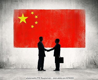 Two figures, both in suits, one with carrying a briefcase, stand in profile shaking hands against a Chinese flag