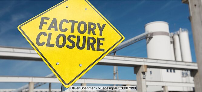 Stock image of a diamond-shaped yellow sign with black "Factory closure" lettering with industrial buildings in the background.