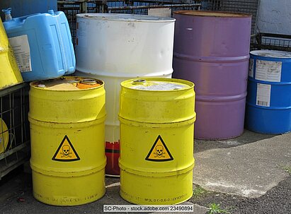 Several steel drums in different sizes and colours. Two of them bear a yellow triangular "skull" hazard sign for toxic substances.  