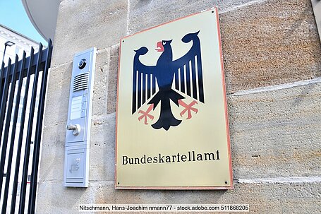 Sign with federal eagle and inscription "Bundeskartellamt" on wall next to doorbell/intercom system and black entrance gate
