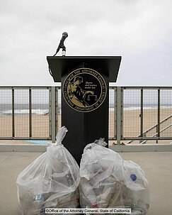 Two bags of plastic waste stand in front of lectern of the california attorney general at the end of the beach
