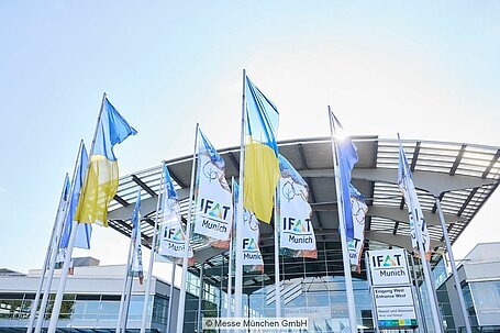 IFAT and Ukrainian flags fly in front of a Messe München building.