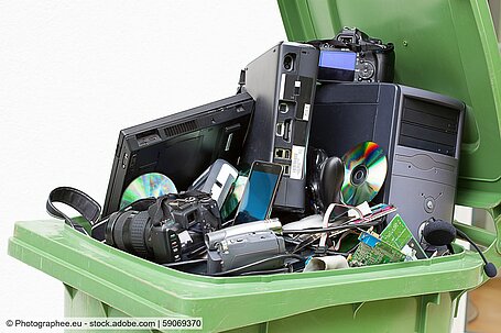 Different types of e-scrap (a PC, a notebook, a smartphone, a headset, cameras, circuit boards, a CD) in an open green waste bin