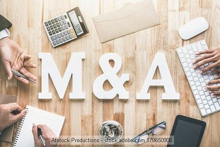 "M & A" as white font on wooden table with a computer, a pocket calculator and office workers' hands