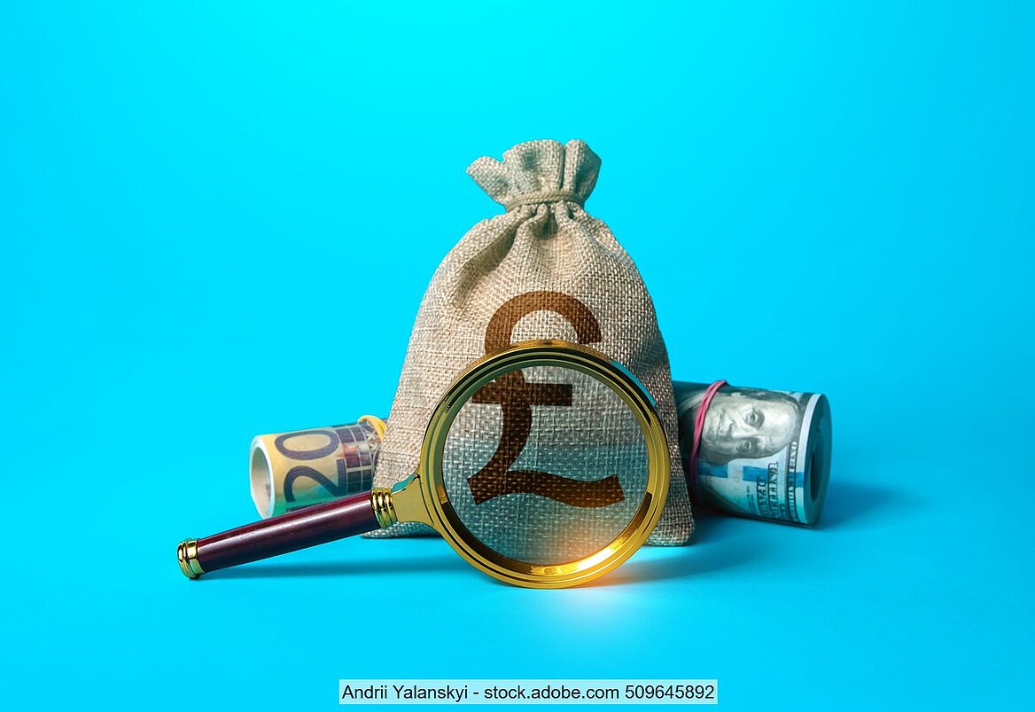 Magnifying glas in front of a sack marked with the £ sign and flanked by rolled dollars and euro notes