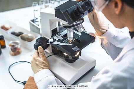 Man in a white lab coat examining sample at microscope, test tubes and other lab equipment in the background (stock photo)