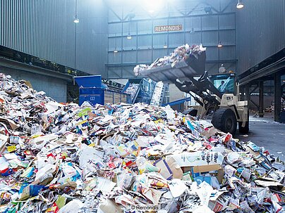 Pile of recovered paper in warehouse, a wheeled loader, conveyor belts and Remondis sign in the background.