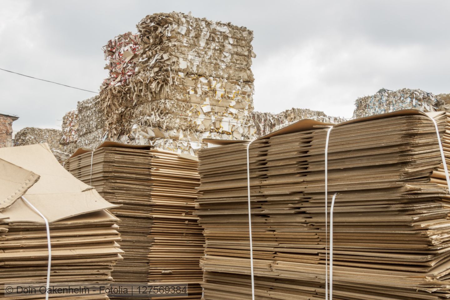 Stacked bales of recovered paper and bundles of flat corrugated board boxes stored in a yard.