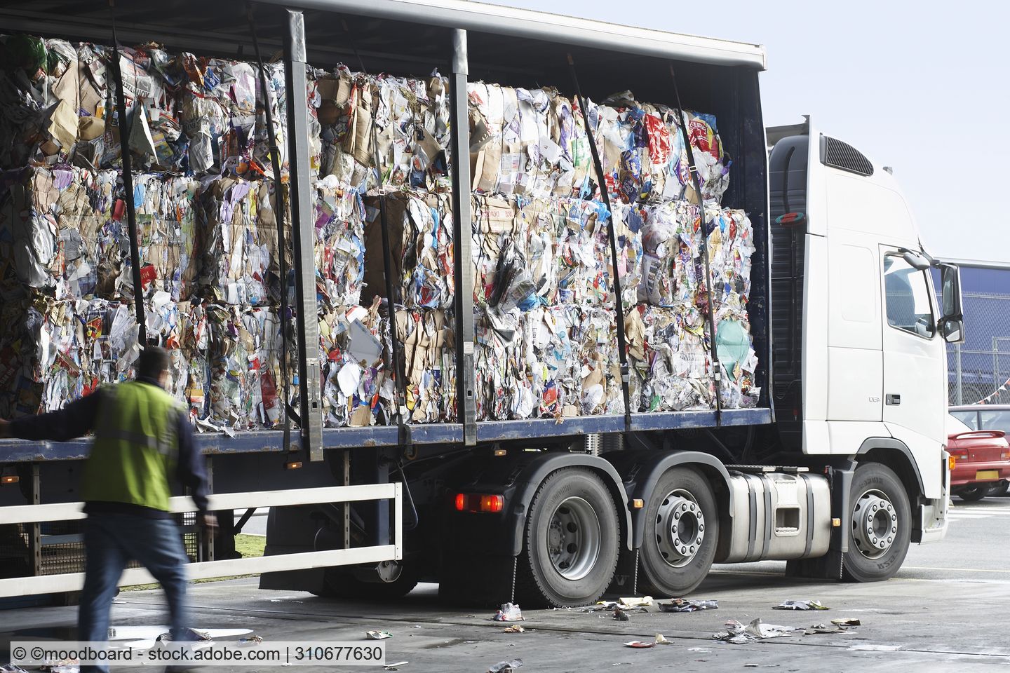 Driver in hi-vis vest inspects stacked bales of waste paper in open curtain-sided trailer