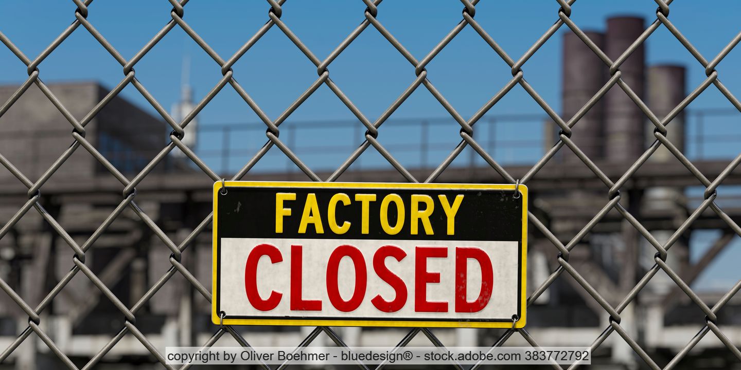 "Factory Closed" sign on a chain-link fence; in the background, factory buildings can be seen.