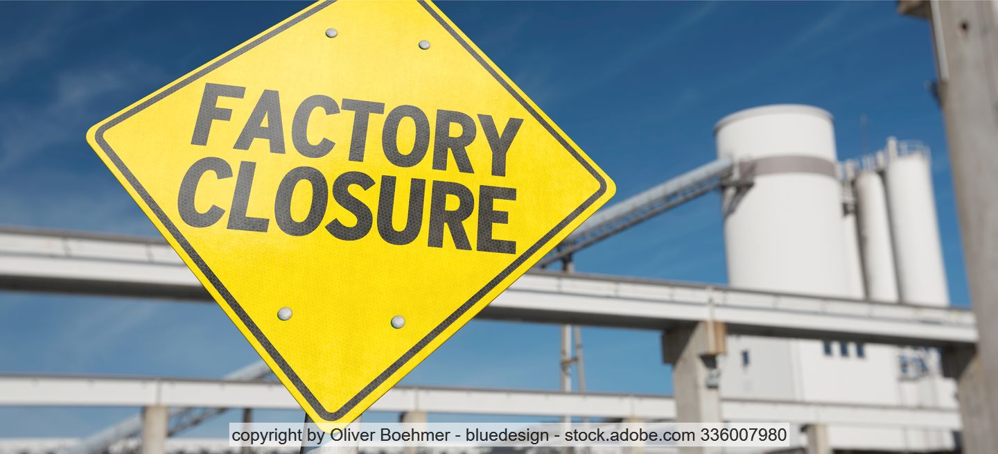 Yellow sign with "Factory closure" printed in black lettering, industrial plants in the background. 