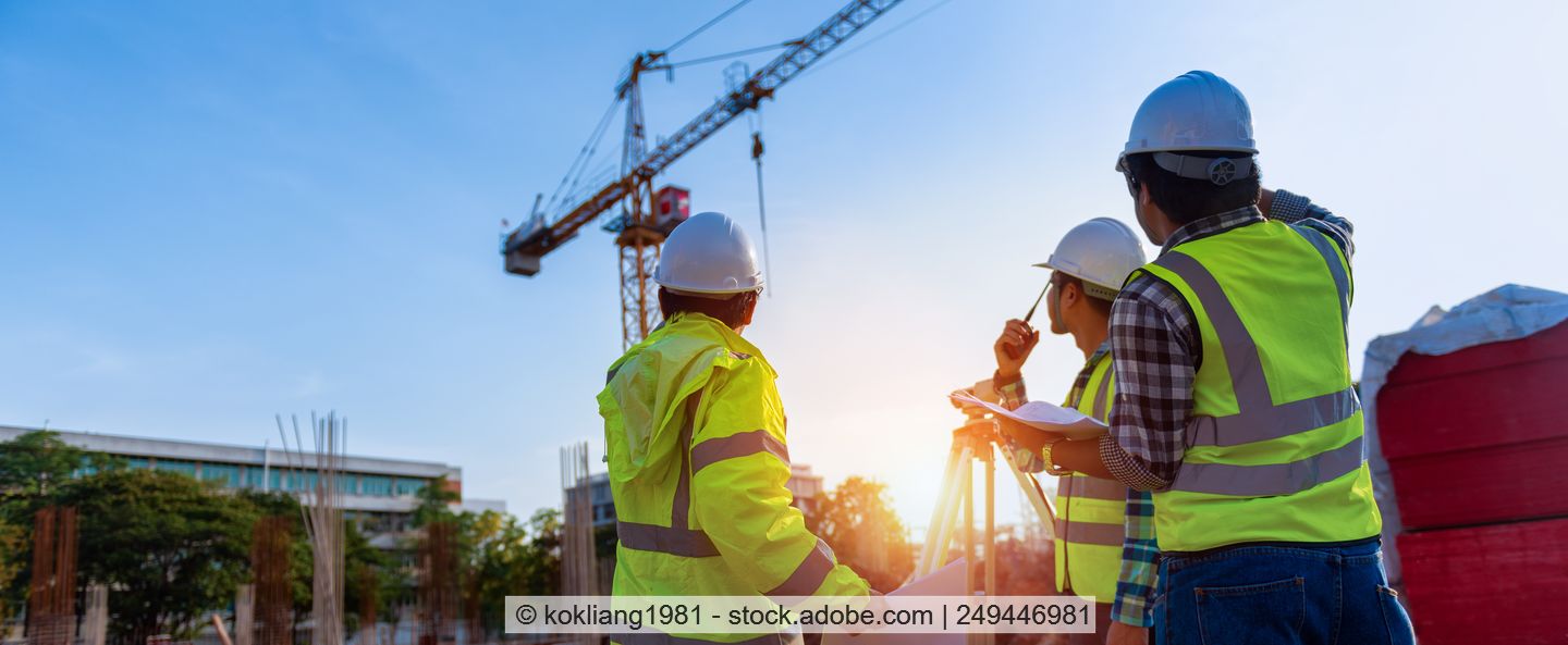 Three men in yellow high-visibility vests stand on a construction site and look up at a crane.