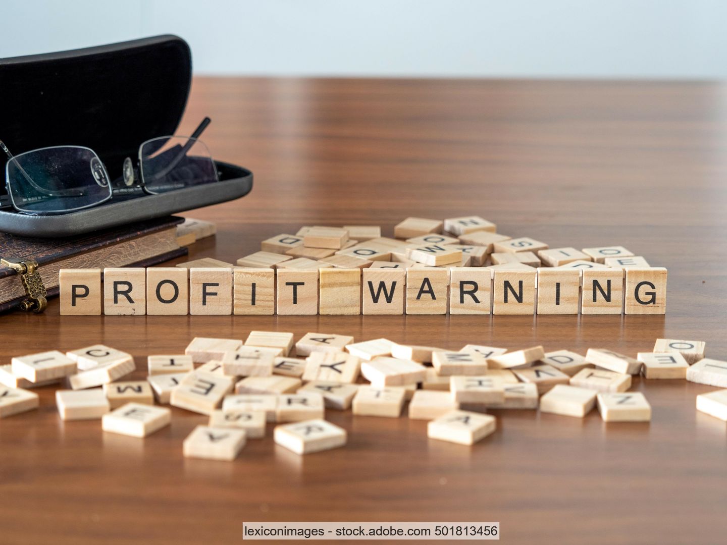 Table surface with wooden tiles with letters on them placed in a row to form the words "Profit Warning", next to them more letter tiles, a pair of glasses and a book. 