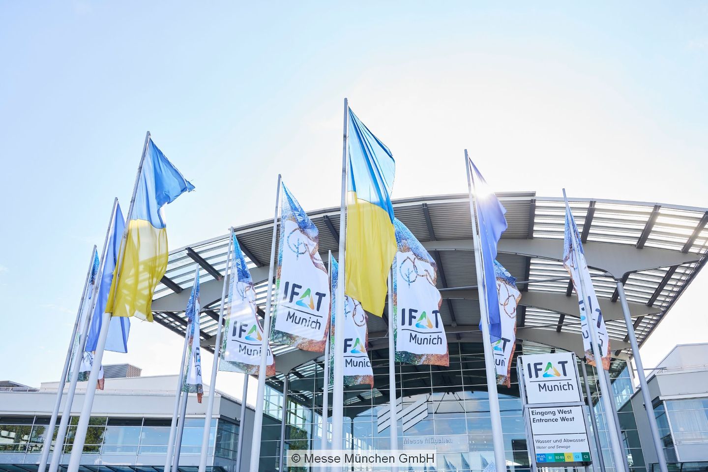 IFAT and Ukrainian flags fly in front of a Messe München building.