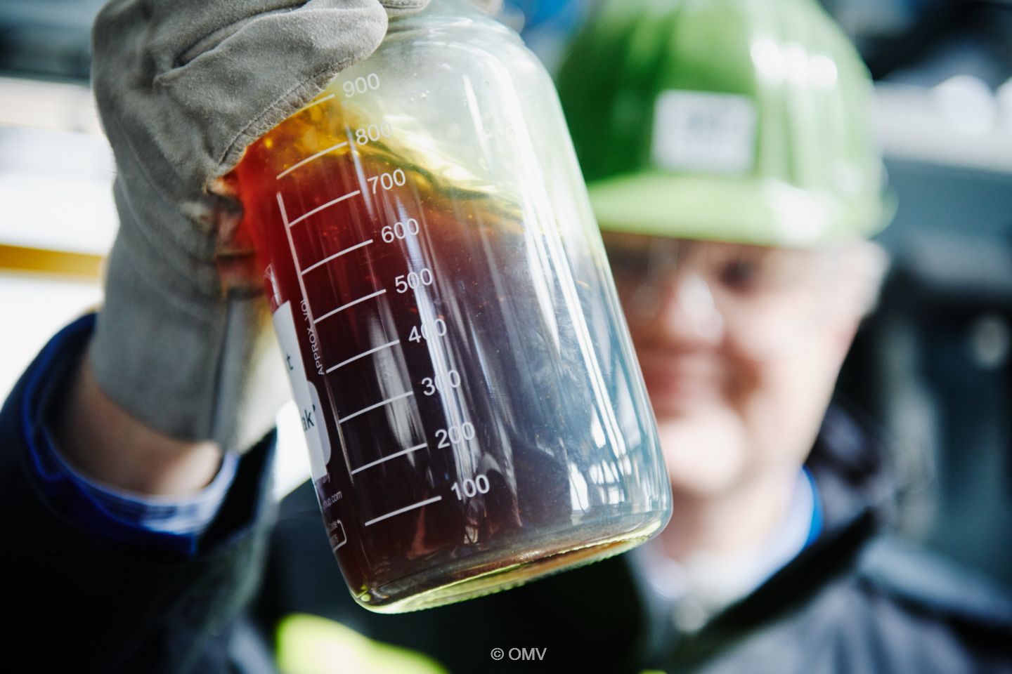 Person wearing a green hard hat holding a glass jar containing a brown liquid.
