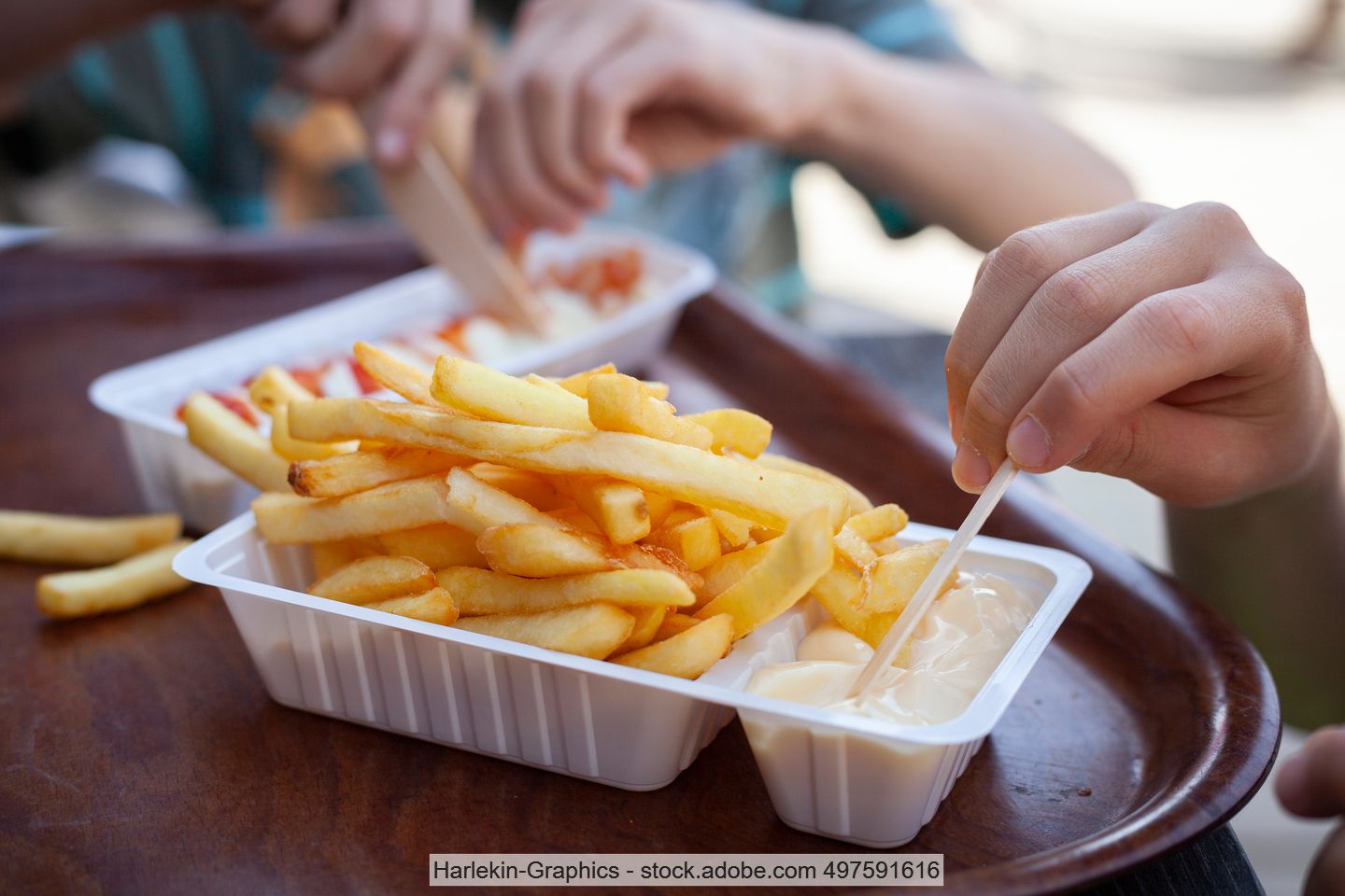 A child's hand pokes a portion of French fries and mayonnaise in a disposable plastic bowl with a chip fork.